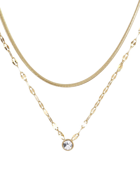 Layered gold necklace with herringbone and link chains, small bezel-set crystal pendant on cream background